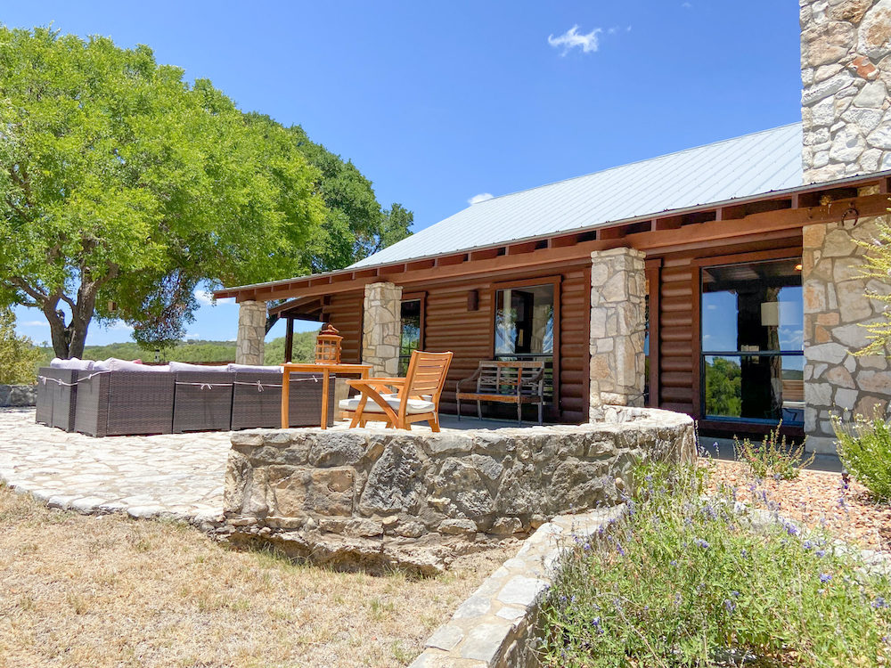 Log-sided lodge with a stone wall, a hot tub and patio seating under a blue sky