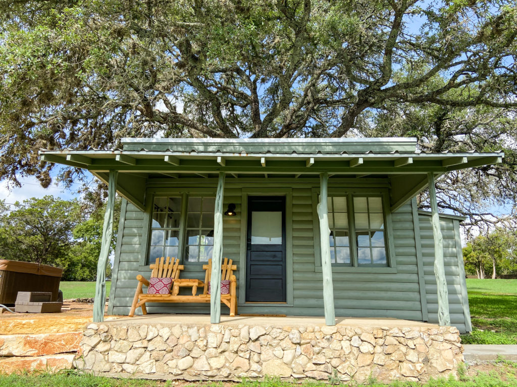 Green cabin with a covered front porch, a wooden bench and a stone foundation beneath a large oak tree