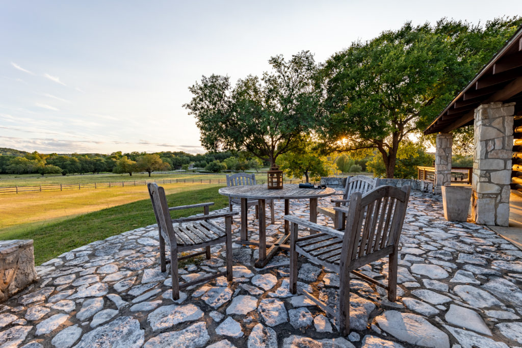 Stone patio with a round table and wooden chairs beside a stone-columned porch, overlooking green pastures and oak trees at sunset