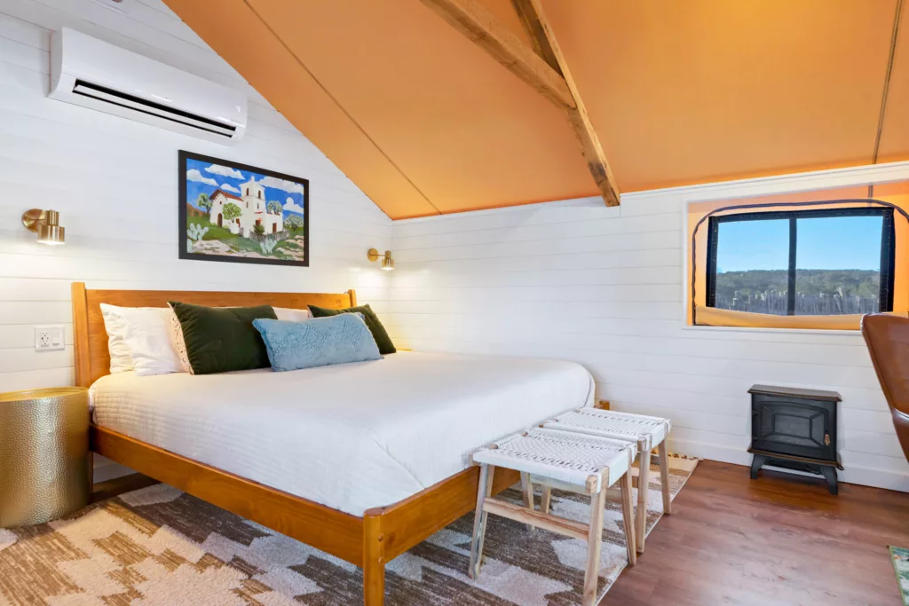 Glamping tent interior with a made bed, a wall-mounted air conditioning unit and a window looking out to the hills