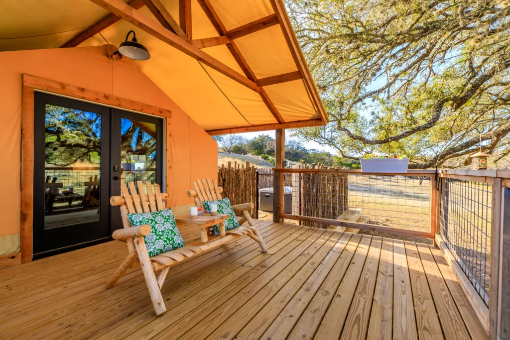 Glamping tent deck with a wooden bench and green pillows beside glass doors, shaded by oak trees