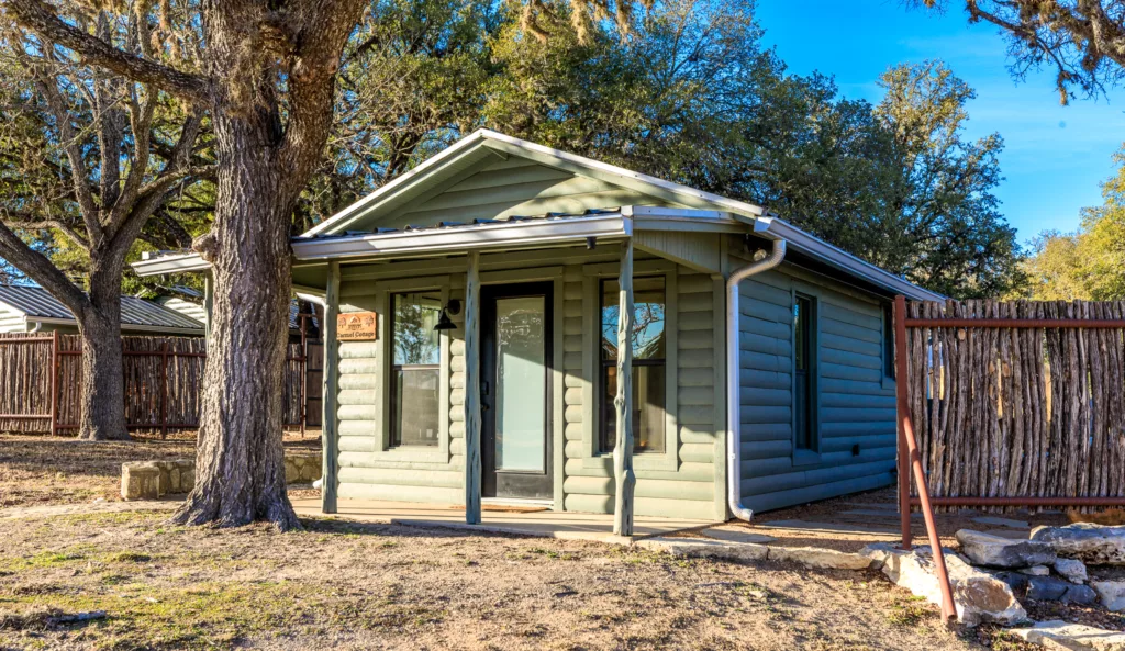 Green cabin with a covered front porch shaded by a large oak tree