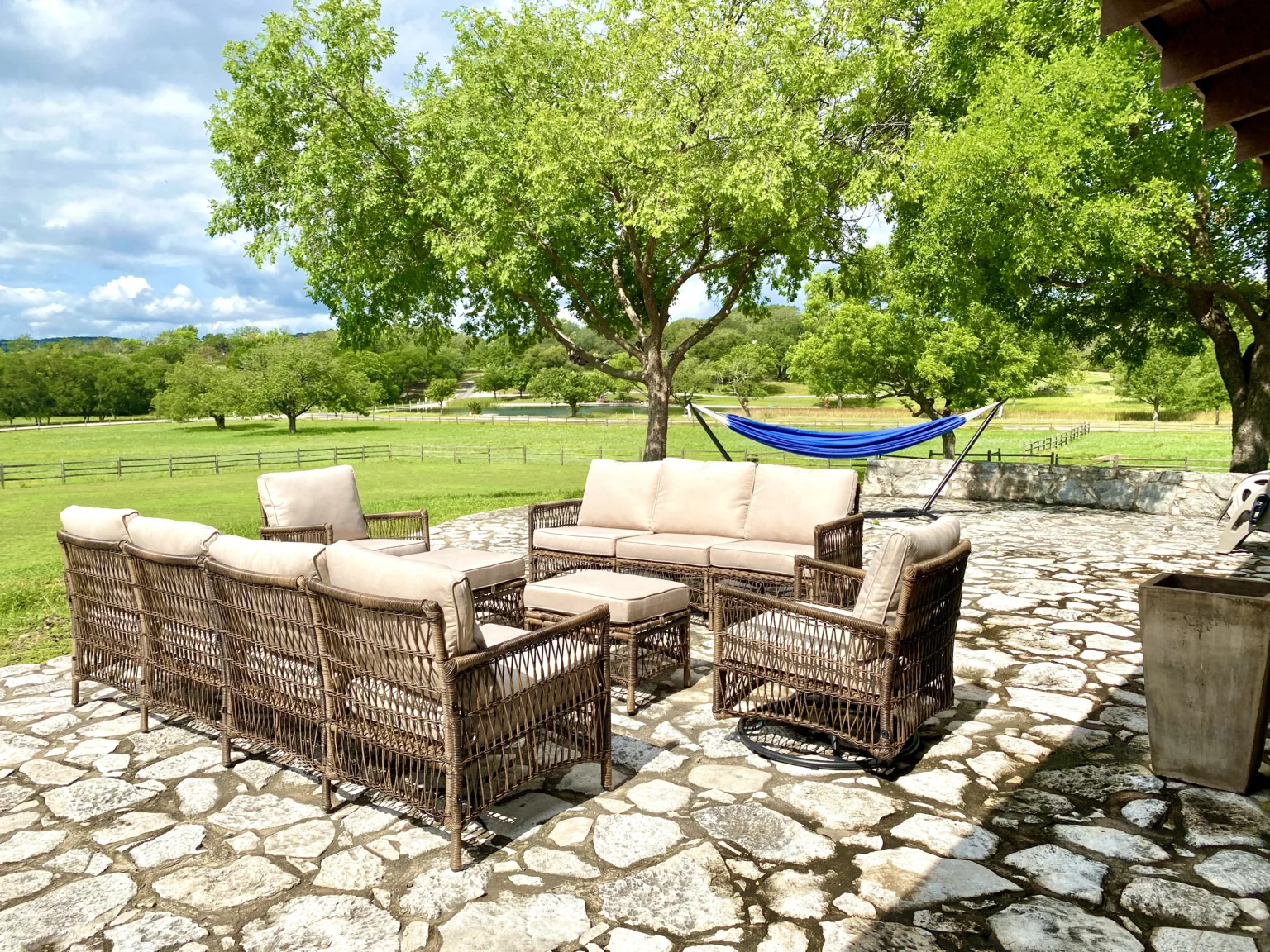 Wicker outdoor sofas and chairs on a flagstone patio beneath shade trees, with a blue hammock and open green pasture beyond