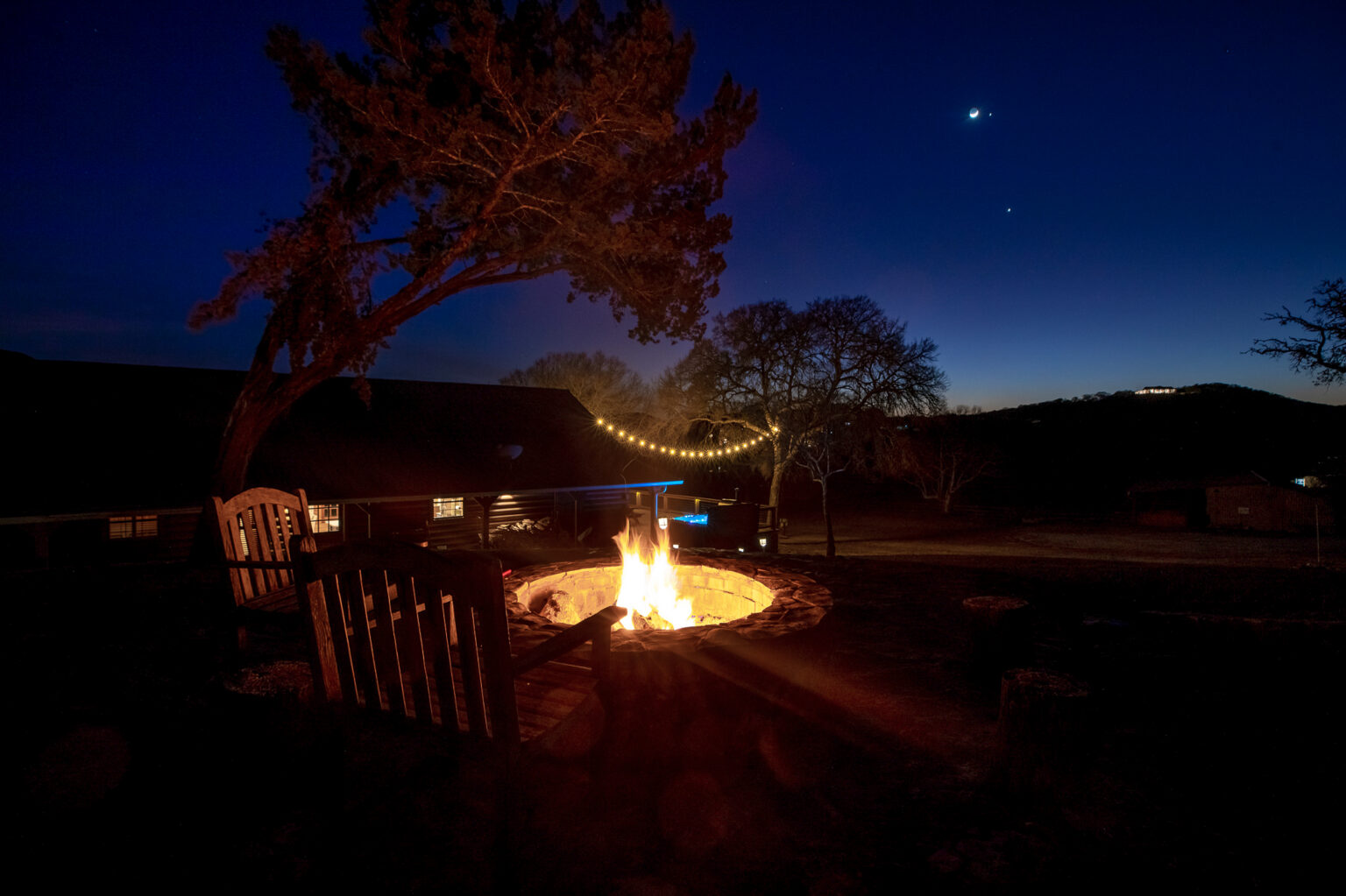 Fire pit glowing at night with wooden Adirondack chairs, string lights and oak trees under a starry sky