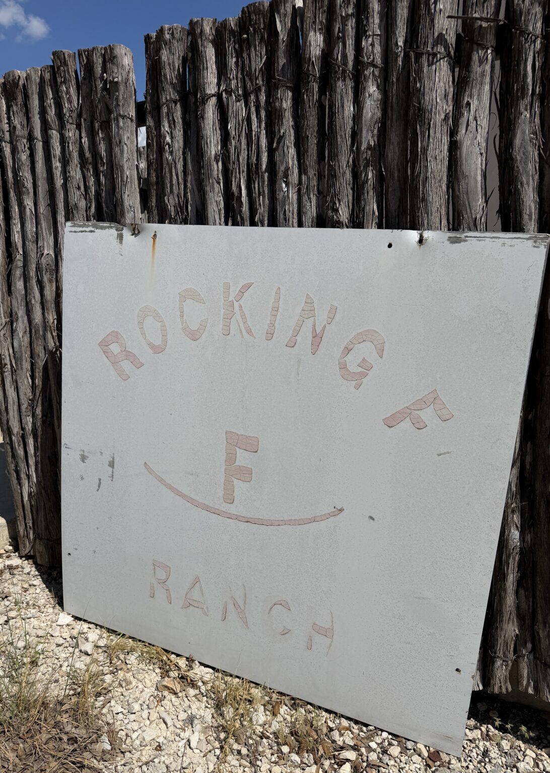 Faded Rocking F Ranch sign with pink mosaic lettering leaning against a weathered cedar post fence under a blue sky