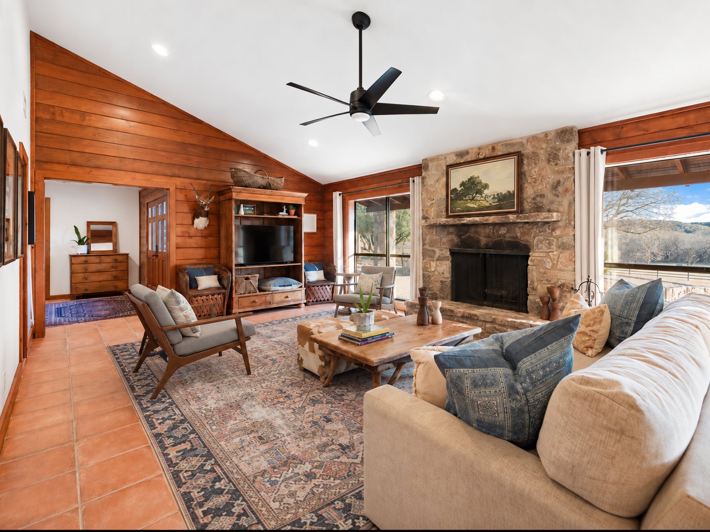 Living room at The Lodge with a vaulted wood-paneled ceiling, a stone fireplace with a landscape painting, a large patterned rug, and a sofa and armchairs around a wooden coffee table