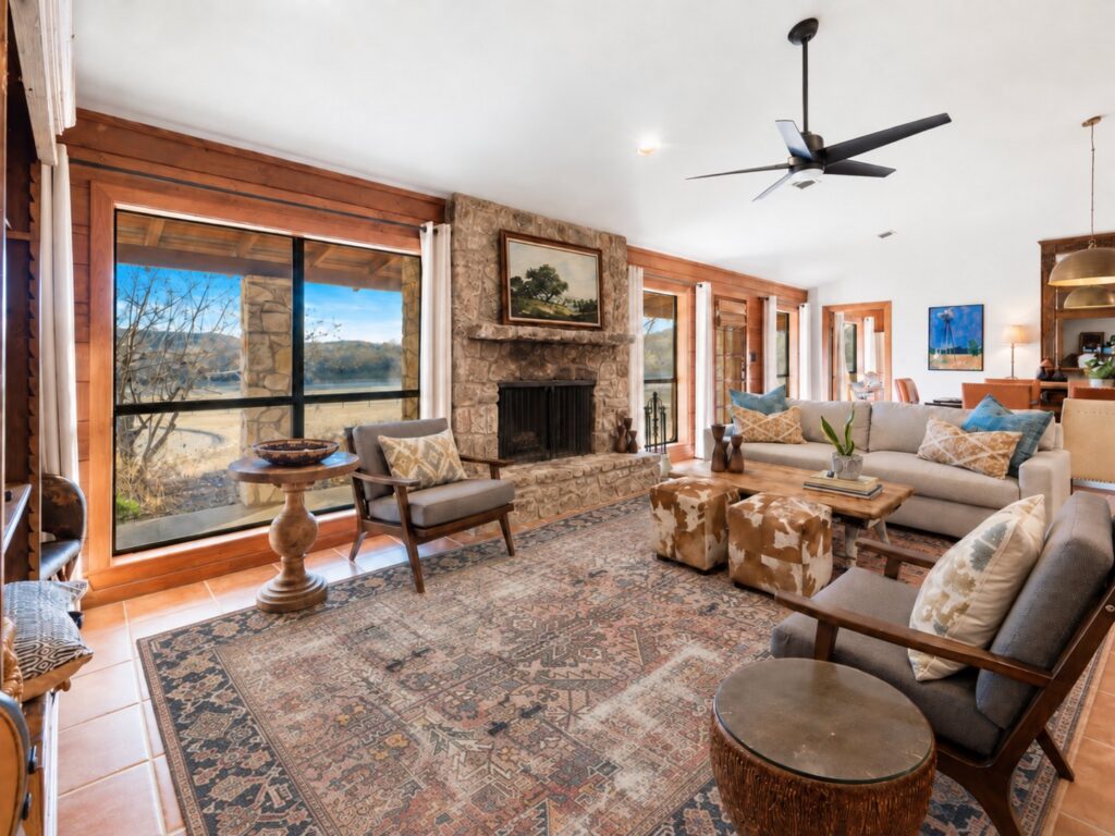 Living room at The Lodge with a stone fireplace and landscape painting, large windows looking out to the Hill Country, a sofa and armchairs around a wooden coffee table with cowhide ottomans, and a dining area beyond
